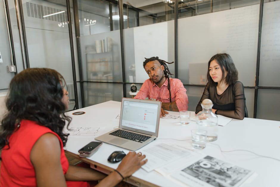 Three professionals engage in an office meeting with a laptop, documents, and discussions.